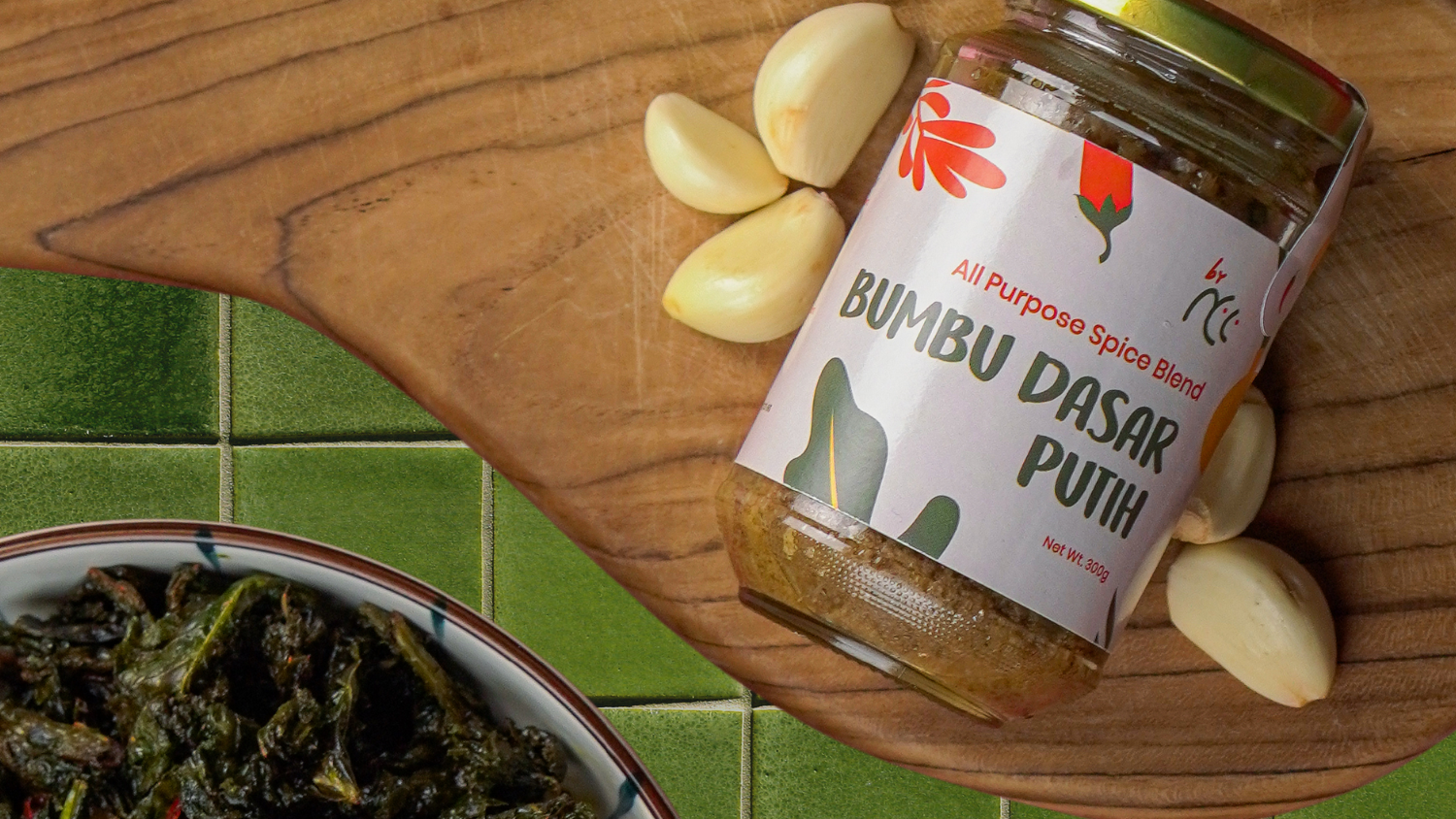 Jar of Bumbu Dasar Putih spice blend by NCC with garlic cloves on wooden board and plate of cooked greens on green tiled surface