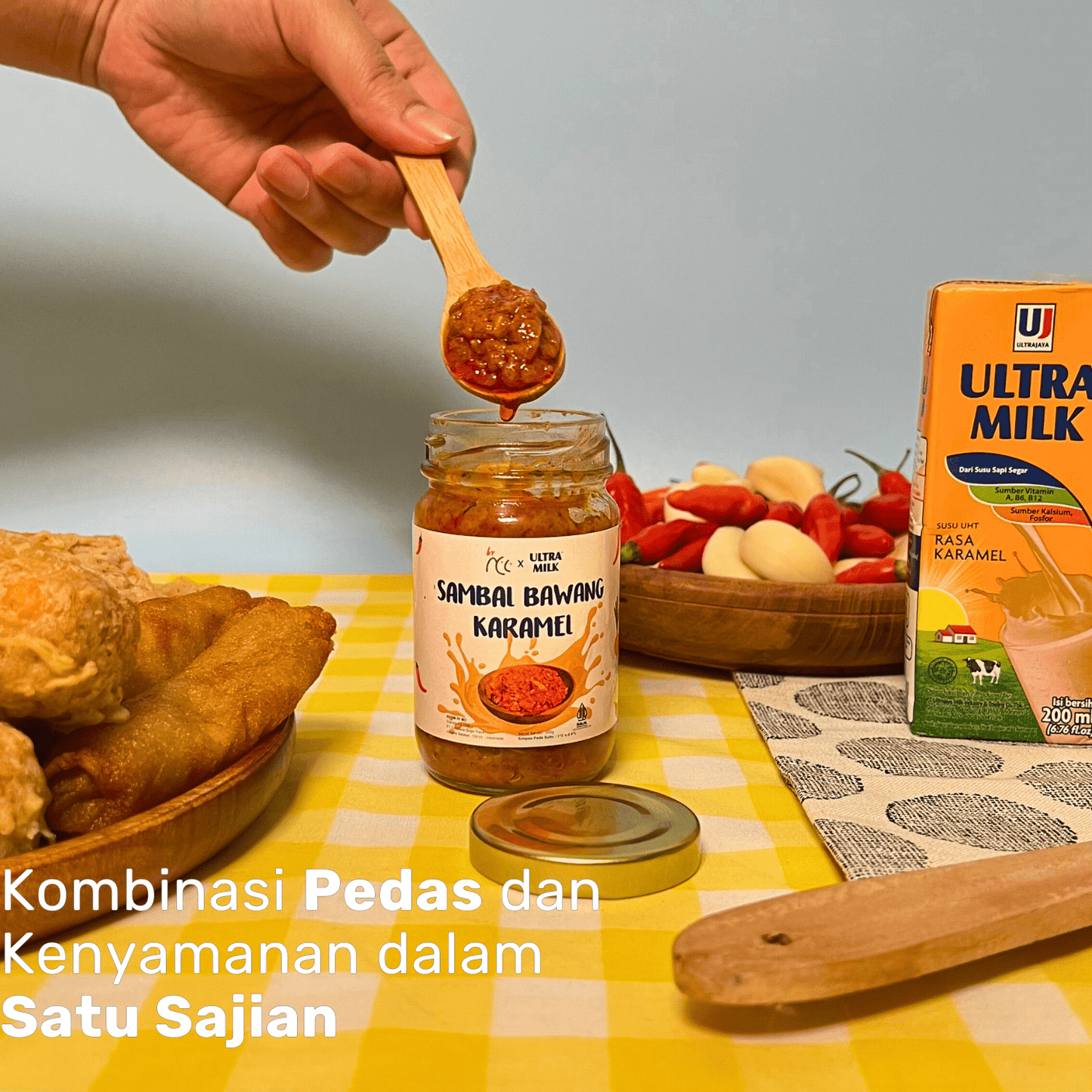 Jar of By NCC x Ultra Milk Sambal Bawang Karamel spicy caramel onion chili sauce with spoon, fried snacks, garlic, chili, and carton of Ultra Milk caramel flavor on table
